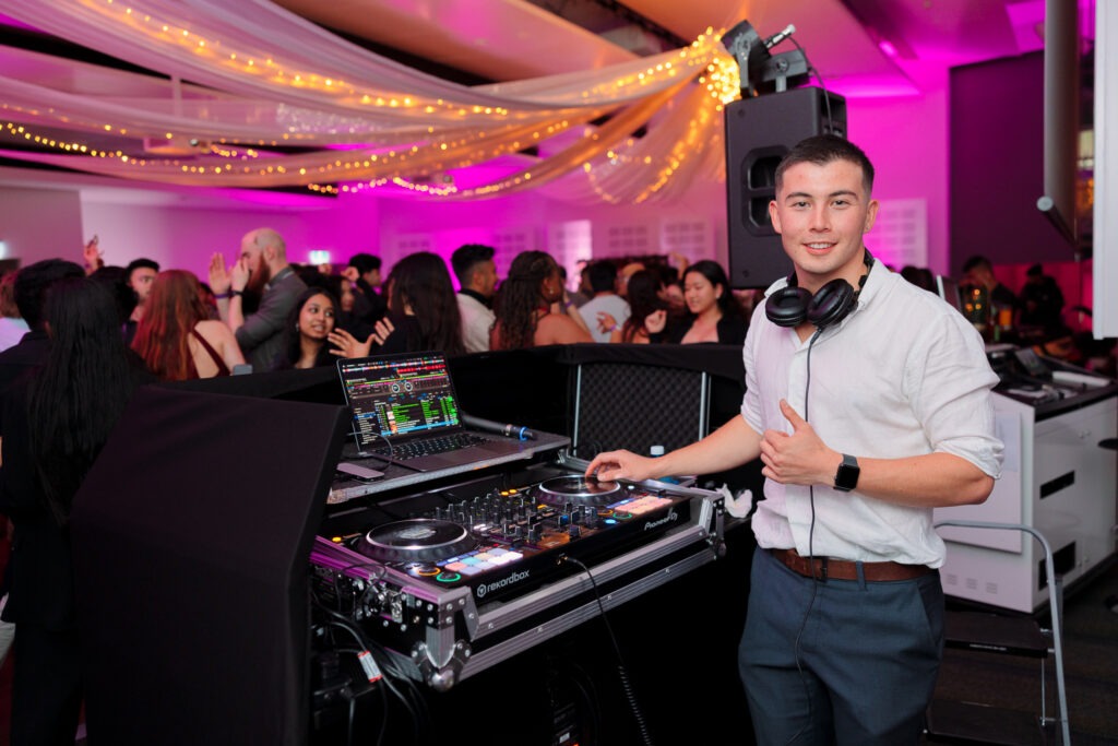 A DJ facing the camera with a colorful themed corporate party and people enjoying in the background, Canberra DJ - 8 til late