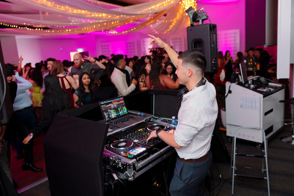 DJ facing the crowd, raising a hand in vibe at a party with people enjoying in the background, Canberra DJ - 8 til late